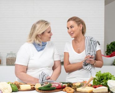 Two women in a kitchen preparing fresh vegetables and cheese with olive oil and spices.