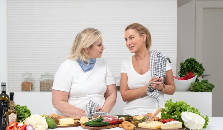 Two women in a kitchen preparing fresh vegetables and cheese with olive oil and spices.