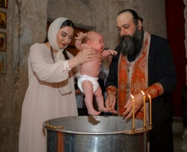 A baby being baptized by a priest and woman in a traditional church setting.