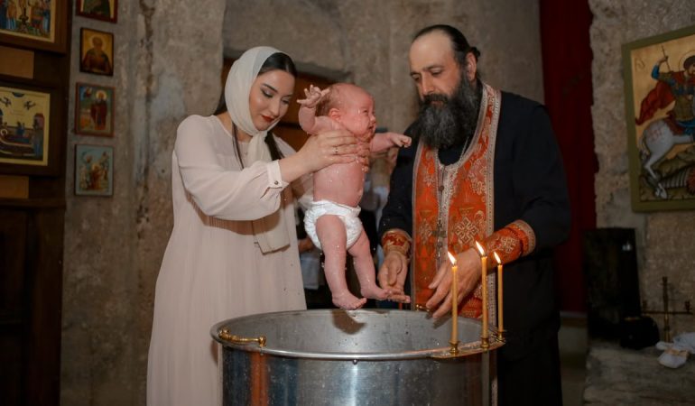 A baby being baptized by a priest and woman in a traditional church setting.